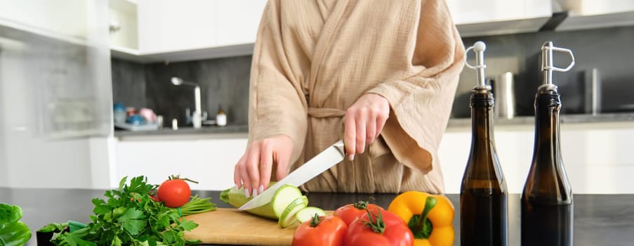 Healthy lifestyle. Young woman in bathrobe preparing food, chopping vegetables, cooking dinner on kitchen counter, standing over white background.