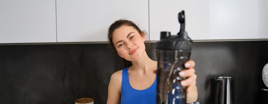 Portrait of beautiful young fitness girl, offering you water bottle to drink after workout, standing in kitchen, smiling at camera.