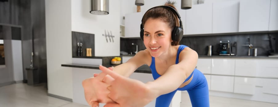 Portrait of beautiful, fit woman in blue leggings and sportsbra, stretching her arms, doing fitness workout, aerobics exercises at home, listening music in wireless headphones.