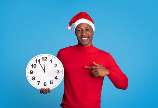 Black Man In Festive Santa Hat Pointing Finger At Clock Standing Against Blue Backdrop In Studio, Counting Down Minutes To Upcoming Year. Excitement Of Christmas And New Year Festivities