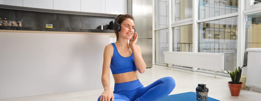 Young sporty athletic fitness trainer instructor woman wear blue tracksuit spend time in home gym use mobile phone headphones listen music. Workout sport concept.