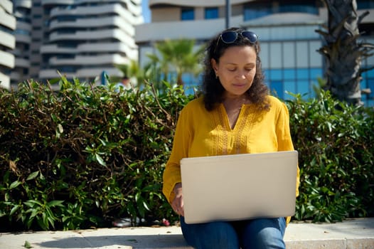 African American adult woman working remotely on laptop, typing text, sitting on a stone bench outdoor. Online business and freelance occupation concept