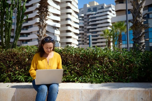 Confident purposeful multi-ethnic female student studying remotely, working on diploma project, sitting on a stone bench against modern high rise buildings background. People. Online education. Career