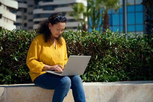 Confident multi-ethnic young adult female student working online on a laptop, sitting on a stone bench against urban buildings background. People. Internet business. Education and career concept