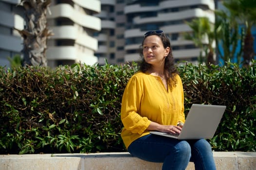 Confident portrait of a female freelancer, entrepreneur using laptop, working online, planning start-up, smiling looking aside while sitting on a stone bench against modern urban buildings background