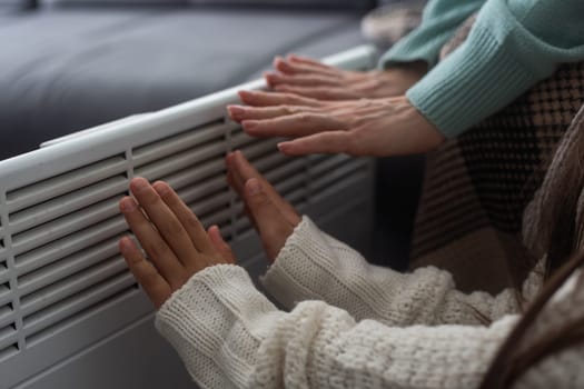 Mother and child warming hands near electric heater at home, closeup