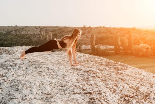 Well looking middle aged woman with long hair, fitness instructor in leggings and tops doing stretching and pilates on the rock near forest. Female fitness yoga routine concept. Healthy lifestyle.