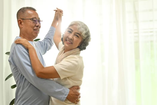 Happy elderly male and female pensioner having fun dancing in living room. Retirement lifestyle concept.