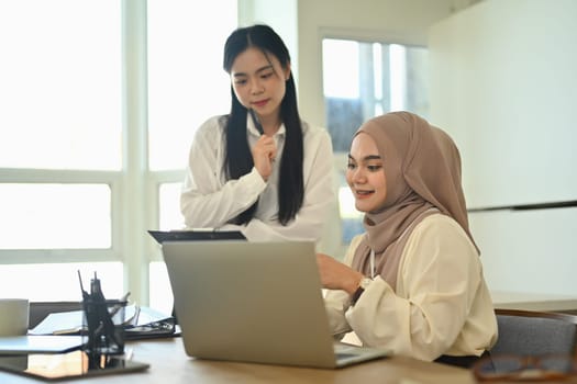 Smiling young Muslim businesswoman using laptop and checking financial document with her colleagues.