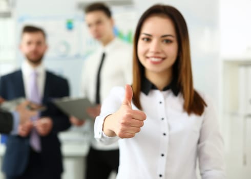 Focus on woman hand showing thumb at camera. Smiling female standing near witty colleagues discussing questionable subject of business in modern building. Company meeting concept. Blurred background