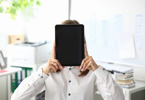 Smart businesswoman sitting at modern workplace and covering face with high-tech digital tablet. Copy space on black blank screen. Advertising concept