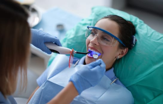 Young woman dentist in protective gloves checking dental seal, fixing photopolymer lamp during stomatology procedure. Dentist specialist visiting patient in dental clinic.