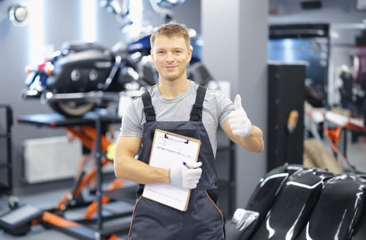 Portrait of young car mechanic holding clipboard and thumb up in car service. Recommendations for high-quality maintenance of cars and motorcycles concept
