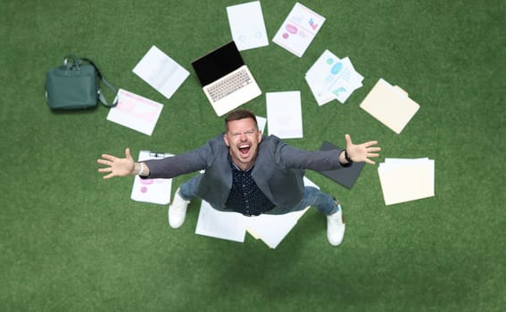 Man in suit throwing papers on green grass and yelling with his hands up top view. Longawaited dismissal from work concept