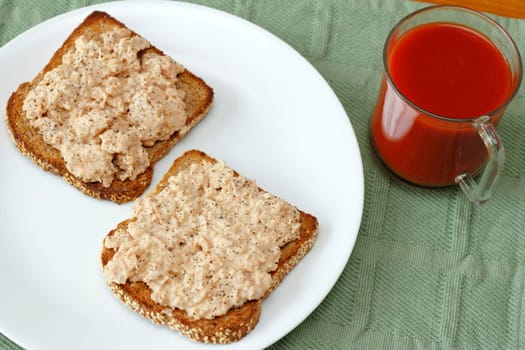Open faced chipotle salmon organic mayonnaise spread on two slices of toasted complete protein sesame sprouted grain bread with a glass mug of tomato vegetable juice.
