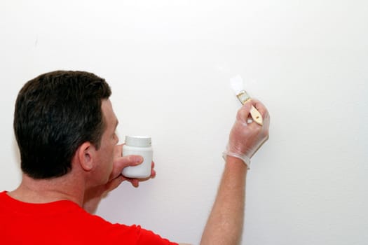 Adult male holding a small glass jar of white touch up paint and wearing a vinyl glove is painting a small mark on a wall.