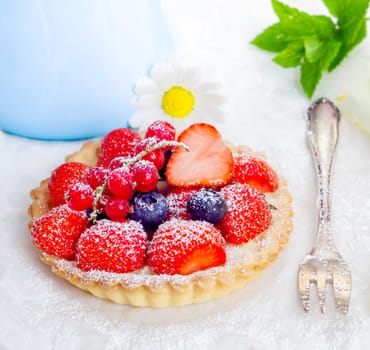 Small individual serving of a fresh whole strawberry, redcurrant and blueberry tart dusted with sugar on a golden crisp crust with a silver cake fork