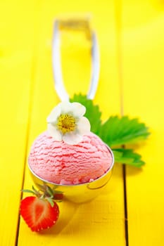 Metal scoop filled with a ball of strawberry icecream garnished with fresh fruit and a flower lying on yellow painted wooden boards. For ice-cream concept look at my portfolio