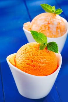 Closeup of a round scoop of colourful creamy orange icecream garnished with mint in boat-shaped porcelain bowls on blue boards