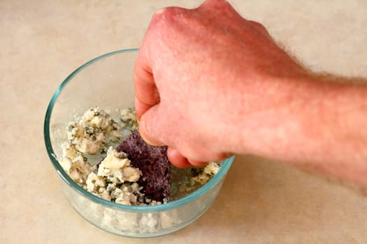 Adult male hand scooping blue cheese crumbles out of a glass bowl and on to an organic blue corn chip.