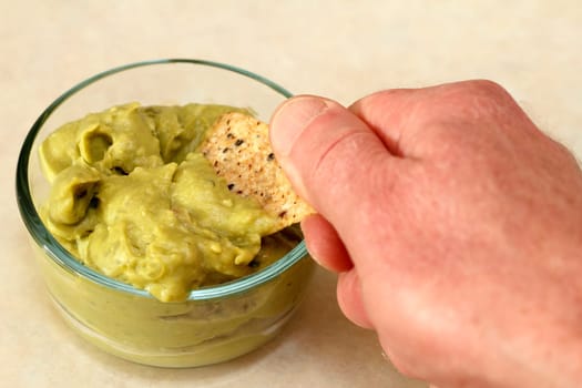 Human caucasian hand dipping organic multi grain mixed olive chip into guacamole dip that&#039 s in a clear glass bowl on a kitchen counter.