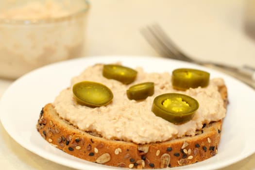Jalapenos top a chipotle salmon spread on one slice of sprouted whole grain bread resting on a plate with a fork and salmon spread bowl in the background.