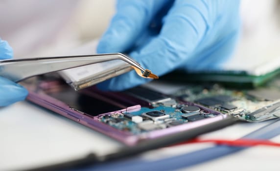 Master in rubber gloves holding microchip over broken phone in workshop. Mobile phone repair concept
