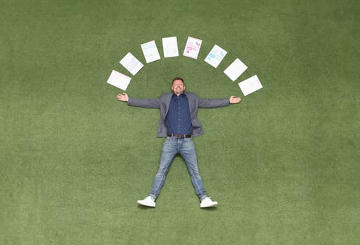 Young businessman lying on green grass with documents around his head top view. Successful business deals concept