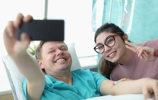 Portrait of smiling young woman and hospital patient taking selfie on mobile phone. Cheerful family picture. Modern clinic room for sick people. Medicine and recovery concept