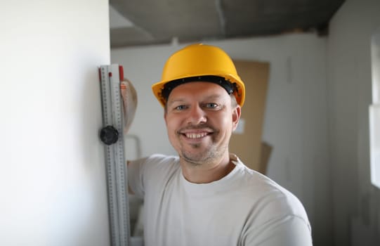 Portrait of cheerful male worker wearing protective yellow helmet checking level. Renovation in apartment. Professional carpenter. Accurate measurement and construction site concept