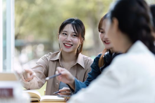 Group of young Asian college students sitting on a bench in a campus relaxation area, talking, sharing ideas, doing homework or tutoring for the exam together.