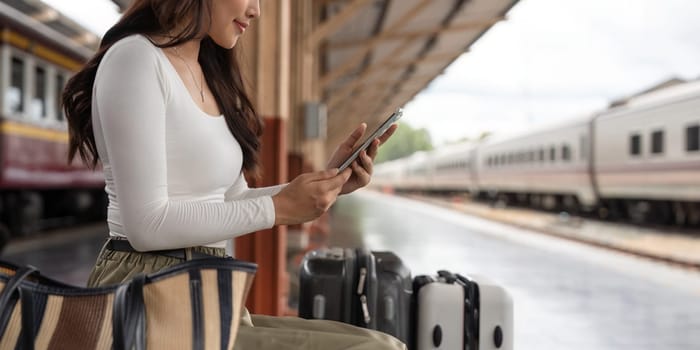 Asian female traveler using her smart phone mobile while waiting for a train at a station.