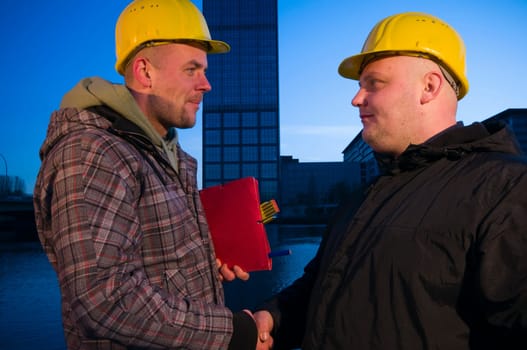 Upper body shot of two men with yellow construction site helmets standing opposite each other and shaking hands and looking into each other's eyes