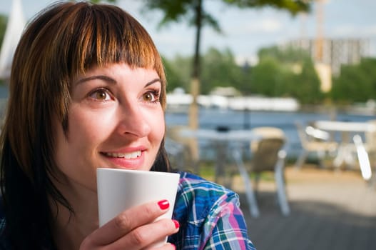 Head and shoulders shot of a young brown-haired woman sitting at a table in a street café and holding a white cup
