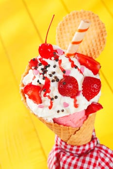 Colourful party ice-cream cone garnished with ripe red strawberries and a spiral wafer biscuit on a cheerful yellow background