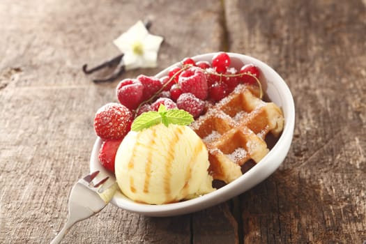 A bowl on a wooden table filled with a golden waffle, icecream and assortment of red berries sprinkled with sugar