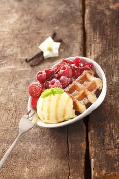 Serving of a crisp golden waffle with icecream, raspberries and redcurrants on an old textured rustic wooden tabletop