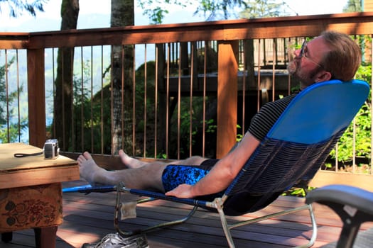 Man relaxing on a lounge chair on an outdoor deck in a forest with his eyes closed.