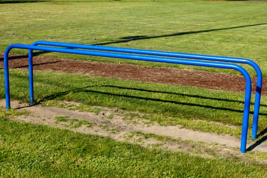 Two blue painted metal parallel bars outside in the grass near a running track on public school grounds under a sunny day in spring.
