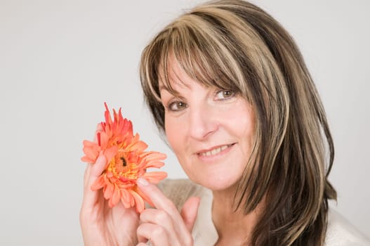 Head and shoulders portrait of middle-aged woman posing with orange-colored flower against neutral bright studio background