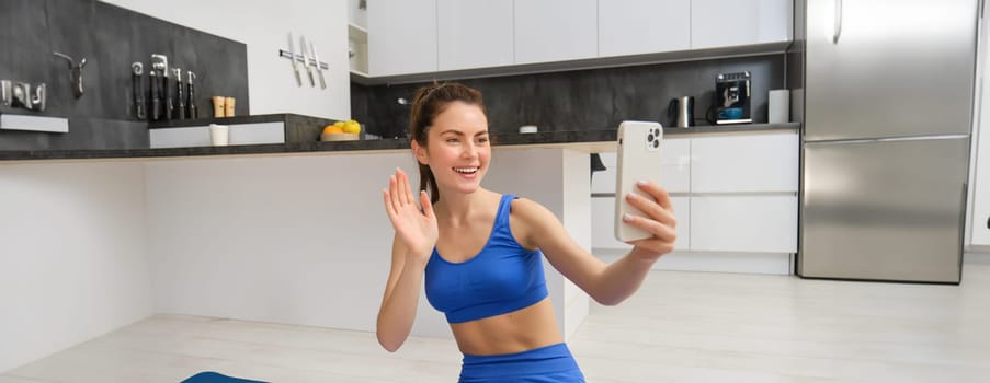 Portrait of young fitness girl, blogger records her workout from home, poses for selfie on smartphone, does yoga on camera, sits in blue sportsbra and leggings.