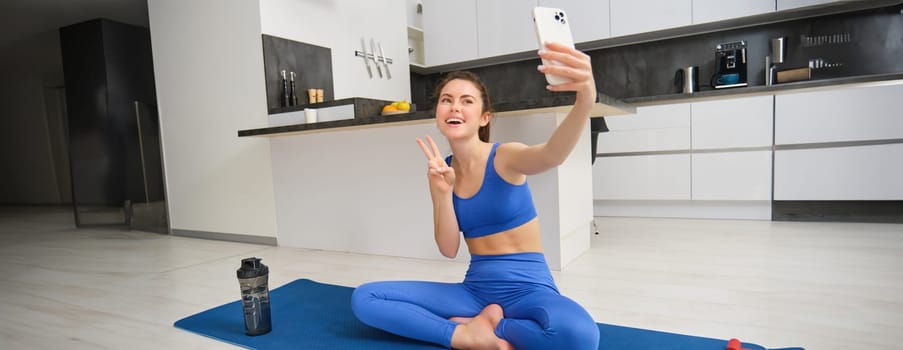 Active young woman, vlogger does sports, records her workout training from home on smartphone camera, posing for selfie inside her house, sits on rubber yoga mat in blue leggings and sportsbra.