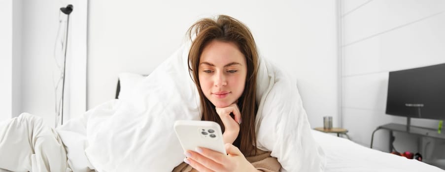 Portrait of cute woman lying in bed and using smartphone, browsing online social media and resting in bedroom in morning.