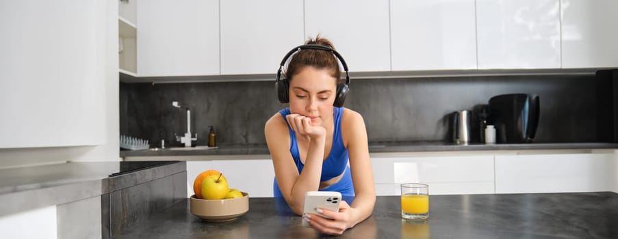 Portrait of fitness girl, standing in kitchen, listening music and looking at smartphone, drinking orange juice from glass, wearing activewear.