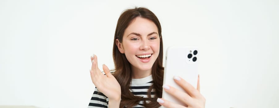 Portrait of young modern girl connects to video chats, talks with friend online using smartphone app, waves at mobile phone camera, white background.