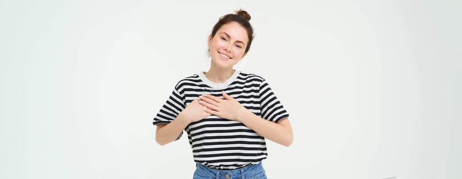 Image of beautiful woman in casual clothes, holds hands on heart and smiles, express gratitude, being thankful, stands over white background.