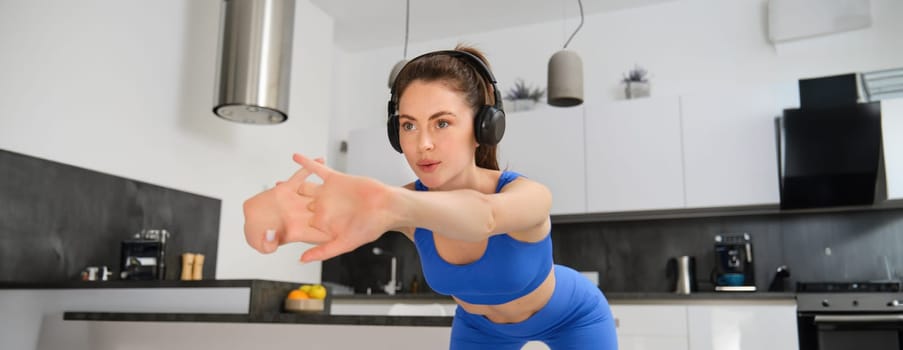 Portrait of fit and healthy woman watching gym workout tutorial at home, stretching body, doing fitness workout in living room, listening music in wireless headphones, doing aerobics exercises.