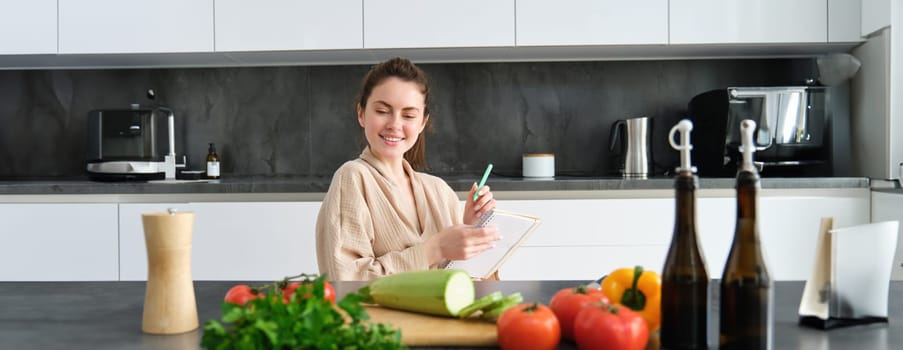 Portrait of woman sitting with groceries, making list for shopping, posing near vegetables, preparing food, cooking in the kitchen.