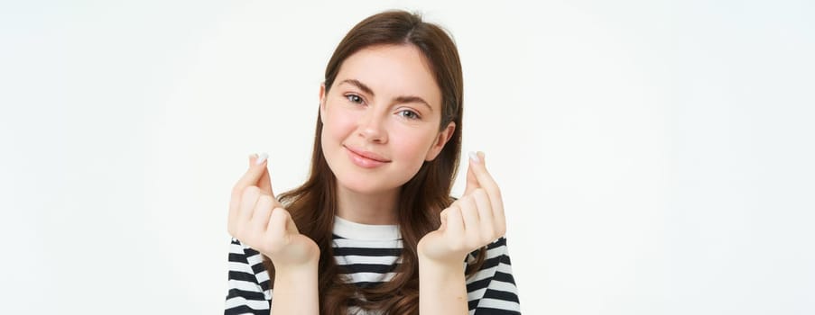 Portrait of beautiful young woman shows finger hearts sign, love and care, likes smth, stands over white studio background.