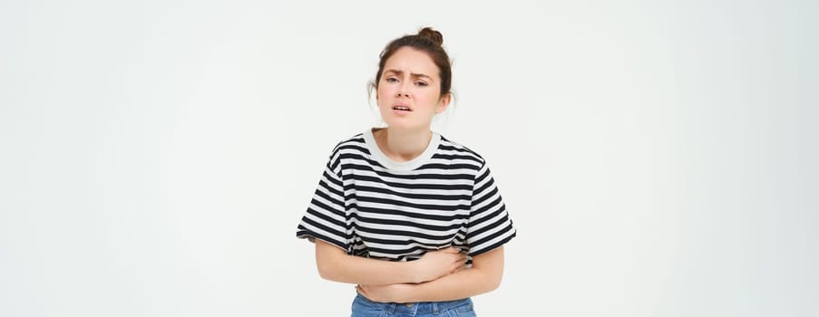 Image of young woman holding hands on her belly, feeling discomfort, menstrual period cramps, needs painkillers, stands over white background.